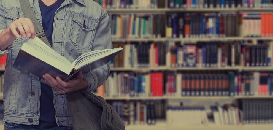 Sie sehen: eine Person mit einem aufgeschlagenen Buch vor einem Bücherregal