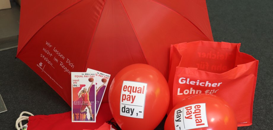 Sie sehen: rote Werbematerialien des Equal Pay Day (Taschen, Luftballons, Regenschirm, Flyer)